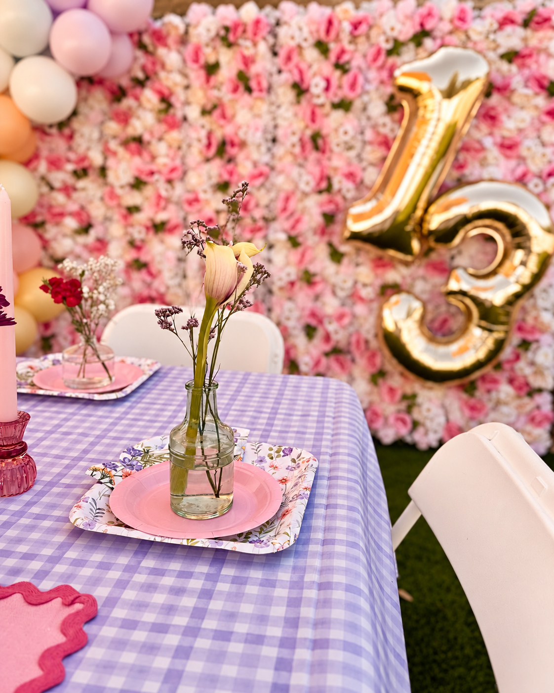 Pink place setting and glass bud vase on a lavender gingham table, with a flower wall and gold balloons behind.